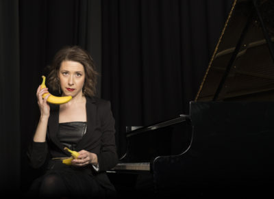 Photograph of Sarah Hagen in front of a piano holding a banana.